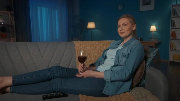 一个年轻的女人正在看电视 喝着红酒 一个女人晚上躺在沙发上 在电视机前休息和放松 — 图库照片
