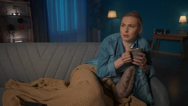 一个年轻的女人躺在沙发上 腿上裹着毛毯 一边喝茶一边看电视 一个女人兴致勃勃地看了一部电影 一部系列片 在剧情结束时 她喝得烂醉如泥 — 图库照片