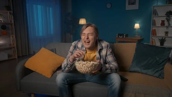 一个男人晚上坐在沙发上 吃爆米花 看电视 男人看喜剧 看喜剧 笑得很开心 — 图库照片