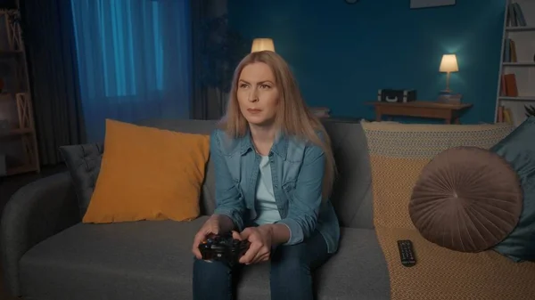 一个女人在晚上坐在家里的沙发上和一个游戏控制器玩一个领事游戏 专心致志的女人按住操纵杆 — 图库照片