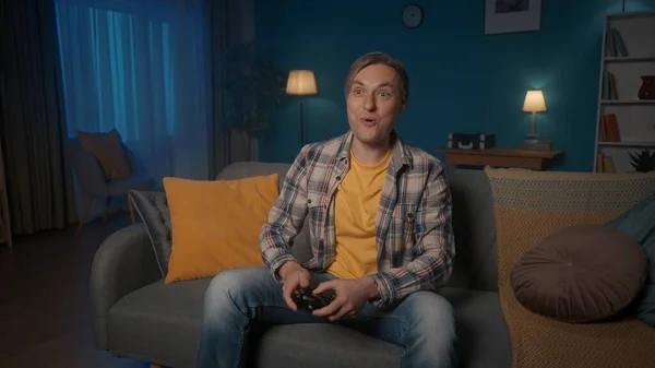 一个男人在晚上坐在家里的沙发上和一个游戏控制器玩一个领事游戏 男人经常按操纵杆 很紧张 — 图库照片
