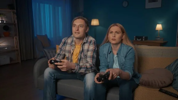 一对年轻夫妇在晚上坐在沙发上与游戏控制器一起玩电子游戏 男人和女人花时间在一起享受游戏 谁会赢游戏概念 — 图库照片