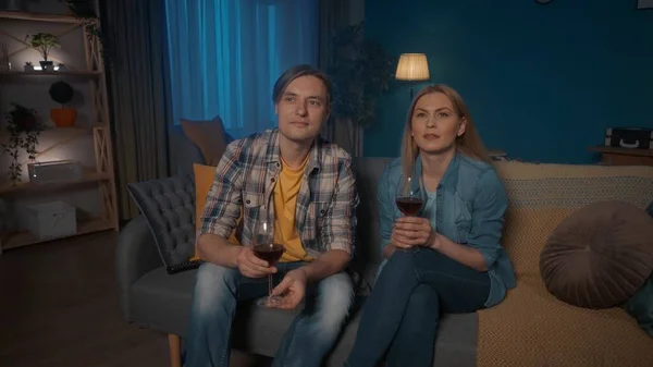 一对年轻夫妇正坐在沙发上 喝着红酒 看电视 一对男女正在饶有兴趣地看一部电影 一部动人的录像 周末休闲概念 — 图库照片