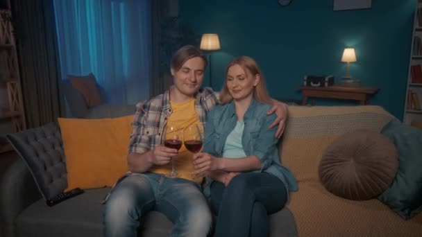 若いカップルは赤ワインのグラスとテレビを見ながらソファに抱きついて座っています 男性と女性が映画 結婚式のビデオ クリンクしてワインを飲んで見ている — ストック動画