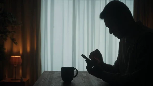 男が携帯電話を持ってテーブルに立っていると コーヒーのカップが近くに立っている 男はテープを通って反転し メッセージを入力し ビデオを見ています 生きている男の暗いシルエット — ストック写真