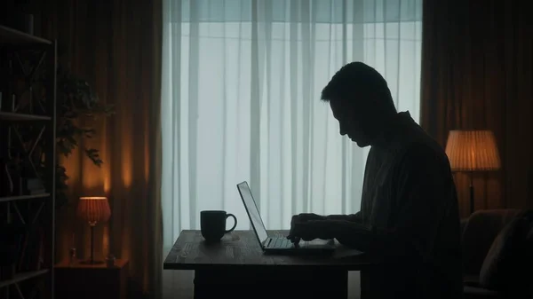 Side view of a dark silhouette of a man at a table in front of a laptop. A man is working on a laptop with a cup of coffee, typing, playing an online game, checking email