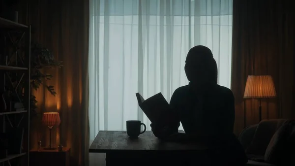 女性は窓の外の部屋でコーヒーと紅茶を片手にテーブルに座っている本を読む 暗いですシルエットの女性とともにオープン本で彼女の手 — ストック写真