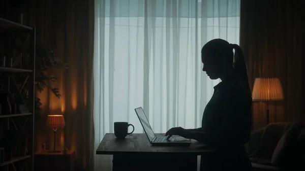在笔记本电脑前的桌子前 一个女人的黑暗轮廓侧视图 一位女士在笔记本电脑上工作 一边喝咖啡 一边玩网络游戏 — 图库照片