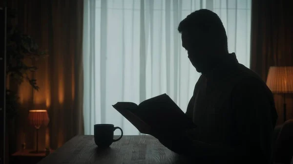 男は窓の外の部屋でコーヒーと紅茶を片手にテーブルに座っている本を読む 暗いシルエットの男とともにオープン本で彼の手を閉じる — ストック写真