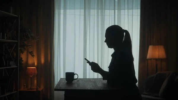 一个女人站在桌旁 手里拿着电话和一杯咖啡 一个女人翻看带子 输入一条信息 看照片 看录像 客厅里一个女人的黑暗轮廓 — 图库照片