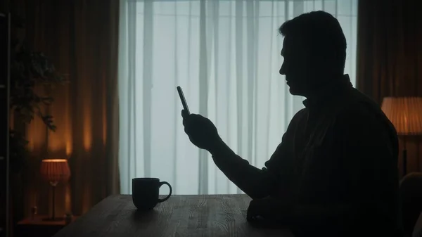 男が携帯電話を持ってテーブルに立っていると コーヒーのカップが近くに立っている 男はテープを通って反転し メッセージを入力し ビデオ通話で話している写真 ビデオを見ています 暗いシルエットのA — ストック写真
