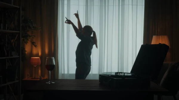 带着乙烯唱片和一杯红酒的复古唱机已经关门了 无忧无虑的女人独自在窗前欢快地跳舞 客厅里跳舞的女人的轮廓 — 图库照片