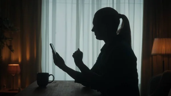 一个女人站在桌旁 手里拿着电话和一杯咖啡 一个女人在视频通话 键入一个消息 看照片 餐厅客厅里一个女人的黑暗轮廓 — 图库照片