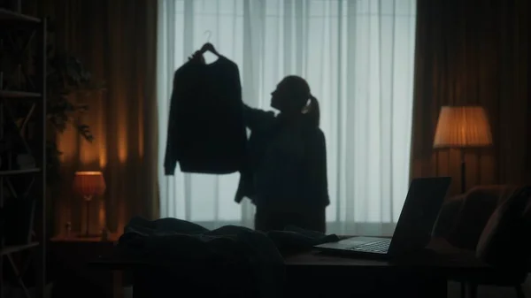 一个年轻的女人选择穿什么 一个女人手里拿着一件衬衫挂在衣架上 仔细检查 以便做出正确的决定 客厅里一个女人的黑暗轮廓 — 图库照片