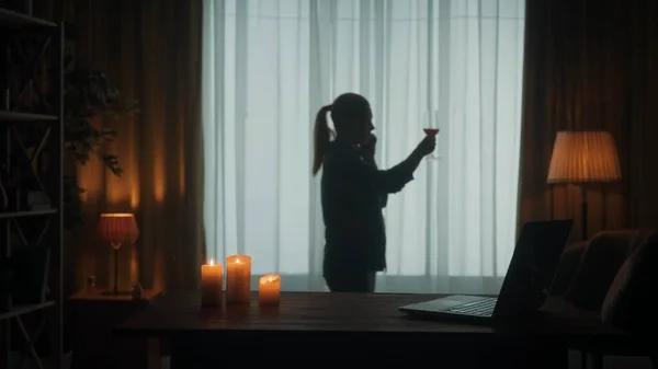 一个女人在讲电话 手里拿着杯酒 桌上放着燃着的蜡烛和笔记本电脑 一位女士正在检查一杯葡萄酒 窗前一个女人模糊的黑暗轮廓 — 图库照片
