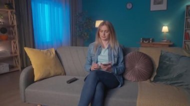 Kanepede oturan bir kadın uzaktan kumandayla televizyonu açar. Bir kadın akıllı bir telefondan akıllı bir ev kontrol uygulaması kullanarak odadaki aydınlatmanın parlaklığını ayarlar. Bir kadın zevk alır.