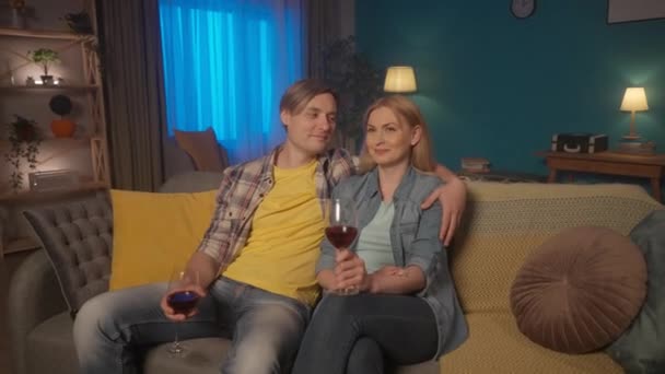 若いカップルは赤ワインのグラスとテレビを見ながらソファに抱きついて座っています 男性と女性が映画 結婚式のビデオを見ています Hdr Bt2020 Hlg材料 — ストック動画