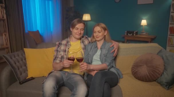 若いカップルは赤ワインのグラスとテレビを見ながらソファに抱きついて座っています 男性と女性は映画 結婚式のビデオ クリンクしてワインを飲んで見ています Hdr Bt2020 Hlg材料 — ストック動画
