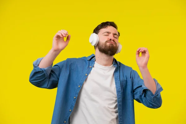 若い大人の髭の男の肖像を気軽に身に着けてヘッドフォンを着用し 音楽に耳を傾け 目を閉じて踊る 黄色の背景に隔離された — ストック写真