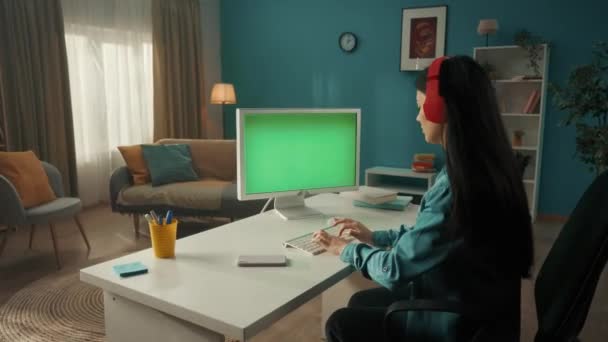 緑の画面のコンピュータで作業しながら ヘッドフォンで音楽を聴くアジアの女性 キーボードで入力する女性 メッセージを書く チャット フリーランス 緑の画面コンピュータモニタ — ストック動画