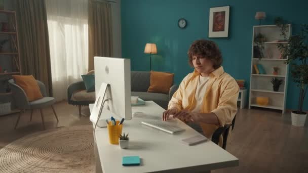 若い男がコンピュータに座っている間にスマートフォンで話す 男は会話を終了し キーボードを入力し メッセージを書き チャット フリーランスになります 若い巻き毛の肖像 — ストック動画