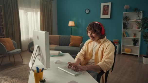 赤のワイヤレスヘッドフォンの若い男がコンピュータに座っています 男は指をスナップし 音楽のビートに合わせて踊り キーボードに入力します スローモーション — ストック動画