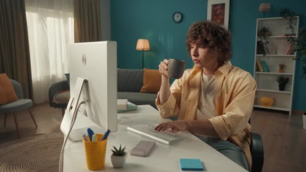 Młody Kręcony Facet Siedzi Przy Komputerze Pije Poranną Kawę Herbatę — Wideo stockowe