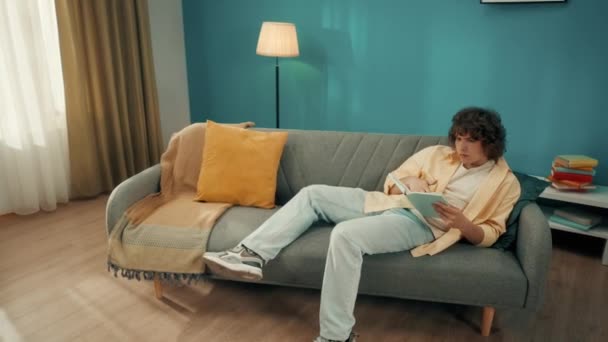 Νεαρός Σγουρομάλλης Διαβάζει Ένα Βιβλίο Στον Καναπέ Μια Ασιάτισσα Δίπλα — Αρχείο Βίντεο