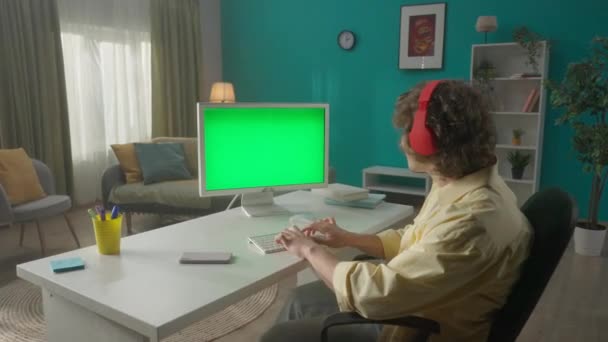 緑の画面のコンピュータで作業しながら 男はヘッドフォンで音楽を聴く 男はキーボードを入力している メッセージを書いて チャット フリーランス 緑の画面コンピュータモニタ — ストック動画