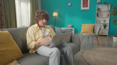 Oturma odasındaki kanepede dizüstü bilgisayarı olan genç bir adamın portresi. Adam video aracılığıyla iletişim kuruyor, selamlaşmak için elini sallıyor, ve başını sallıyor,