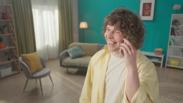 一个年轻人在智能手机上说话 用食指指着家具 告诉对话者他的住房情况 客厅里一个带着电话的卷发男子的画像 — 图库视频影像