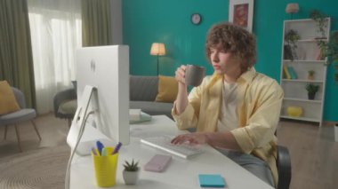 Genç, kıvırcık bir adam bilgisayarın başına oturur ve sabah kahvesini ve çayını içer. Klavyede yazan, e-posta yazan, mesaj bırakan, sohbet eden, online çalışan, serbest çalışan bir adam. Ağır çekim. HDR BT2020 HLG Malzemesi