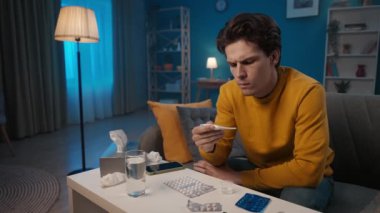 A man measures the temperature with a thermometer, sitting at home on the couch, in front of a table with medicines. A sick man, having measured the temperature, drinks syrup, antipyretic. Home