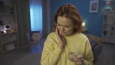 Young attractive woman drinks water with ice while standing in the living room. The woman grabs her cheek, with a suffering expression, feels pain in her teeth. Tooth sensitivity, toothache. Home