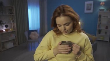 A woman holds a cup of hot tea in her hands, blows on it and drinks. Woman clutches her cheek, rubbing it, suffers from toothache due to tooth sensitivity. Tooth sensitivity, toothache. Home medicine