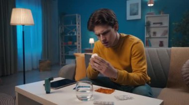 Bir adam oturma odasındaki bir kanepede oturuyor, önünde çeşitli ilaçlar var. Bir adam bir şişe hap alıyor, adını okuyor ve ilaçların talimatlarını çalışmak için tableti kullanıyor.