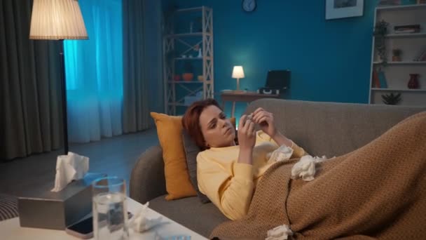病気の女性はリビングルームのソファーに横たわり 温度を測定します 女性は驚きの結果を見て 彼女の鼻を紙のハンカチに吹き込む 女性は高いから苦しむ — ストック動画