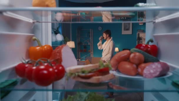 打开冰箱 架子上放蔬菜 香肠和三明治 一个穿着浴衣的男人站在那里喝咖啡 满意地看着产品 点点头 从里面看 — 图库视频影像