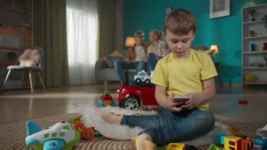 Annem ve babam kanepede oturup konuşuyorlar. Tatlı çocuk oyuncaklarla çevrili yerde oturuyor. Elinde bir akıllı telefon tutuyor ve bir şeyler izliyor, oynuyor. Aile birlikte oturma odasında..