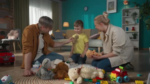 若い大人の両親と子供は一緒に時間を過ごしています 子供は部屋の床に座り たくさんの玩具に囲まれていました 彼らは男の子に見て 話して笑顔でおもちゃを与えています — ストック動画