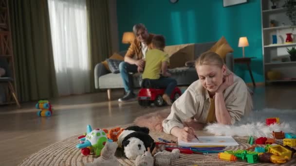 リビングのファミリーシーン 父親はソファーに座って息子と話し 踊り始める ママは床に横たわり 色鉛筆でアルバムに何かを描いています — ストック動画