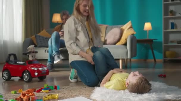 スマートフォンでソファーに座っている大人のお父さん 母と息子は床に座っている 母親は子供をくすぐり始めました 彼らは笑顔で 一緒に時間を楽しんでいる スローモーション — ストック動画