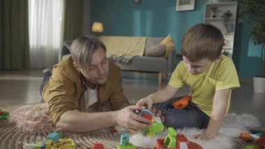 Yetişkin bir baba ve çocuğunun oturma odasında yerde yatarken çekilmiş yakın plan bir fotoğrafı. Odaklanmış çocuk elinde otomatik bir tornavida tutuyor ve babası oyuncak bir uçağı tamir etmesine yardım ediyor. Baba ve oğul mutlu.