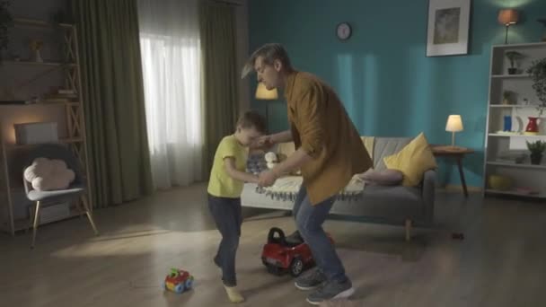 年轻的成年父亲和他的小儿子手牵着手 在客厅里愉快地跳舞 爸爸抱着他的儿子 把他扶起来 家人在一起玩得开心 — 图库视频影像