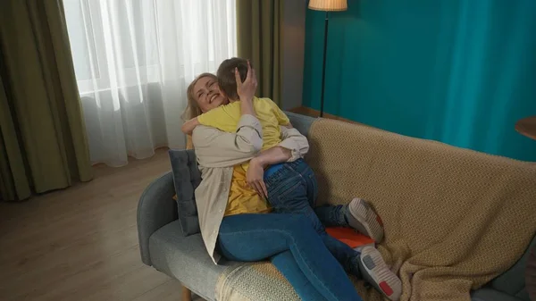 Candid family scene in the living room. Young adult mom sitting on the couch and holding her son, hugging and kissing each other. Happy relationships mother and son.
