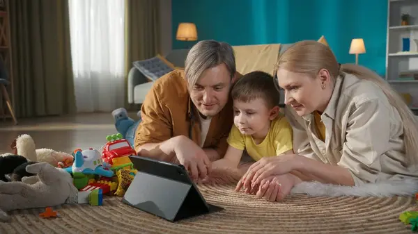 Family spending time together at home. Young parents and their cute son laying on the floor in living room and watching tablet. Smiling mom and dad poiting at the screen and talking to their child