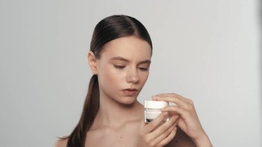 Kozmetik ürünler taşıyan at kuyruklu ve doğal makyajlı esmer bir kadının portresi. Kadın bir krema kavanozunu açar, kameradan uzağa bakar. Beyaz arka planda izole edilmiş. Cilt bakımı