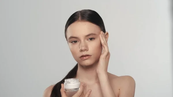 化粧品を保有する慎重なブルネットモデルの肖像 彼女は開いた瓶を持ち カメラを見ている彼女の顔にクリームを塗ります 白い背景に孤立した スキンケア広告 — ストック写真