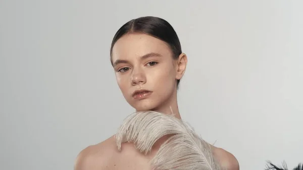 魅力的なブルネットの女性の肖像画 白い羽で彼女の顔に触れるカメラを見ているモデルのクローズアップショット 白い背景に孤立した スキンケア広告コンセプト — ストック写真