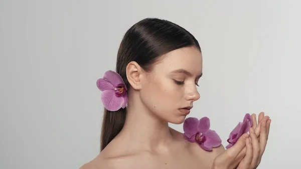 Portrait of a appealing brunette female model. Close up shot of a woman with ponytail holding an orchid flower in her hand and looking at it. Organic skincare advertisement concept.